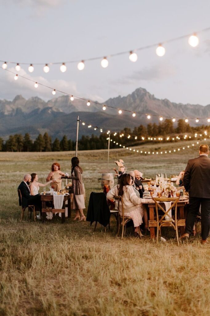 Outdoor wedding reception with guests under string lights, set against mountain backdrop at dusk.