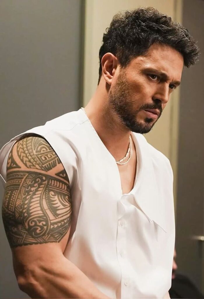 Man with a tattooed arm in a white shirt, looking focused and thoughtful indoors.