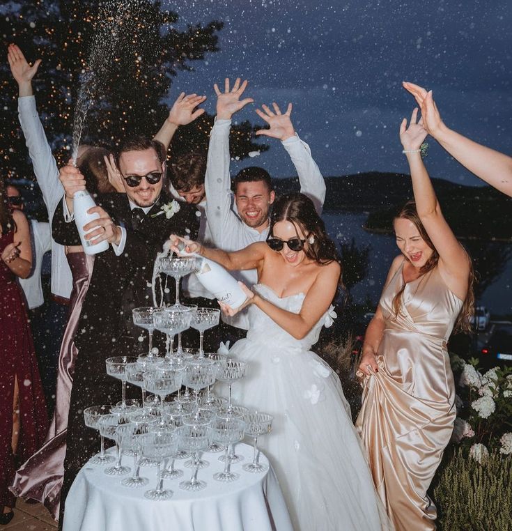 Bride and groom celebrate with champagne cascade at outdoor wedding night party, surrounded by friends.