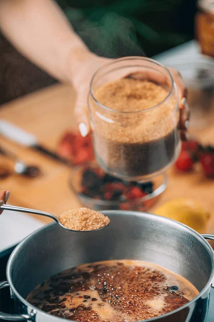 Adding brown sugar to a boiling mixture in a pot, with berries and ingredients in the background.