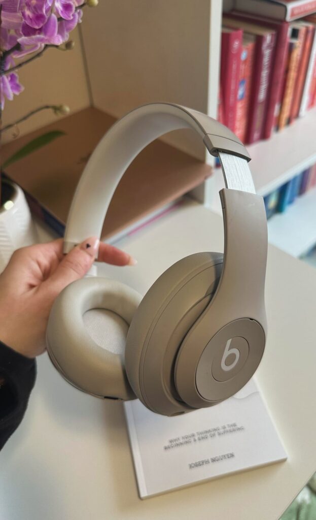 Gray Beats headphones held by a hand, with bookshelf and purple flowers in the background.