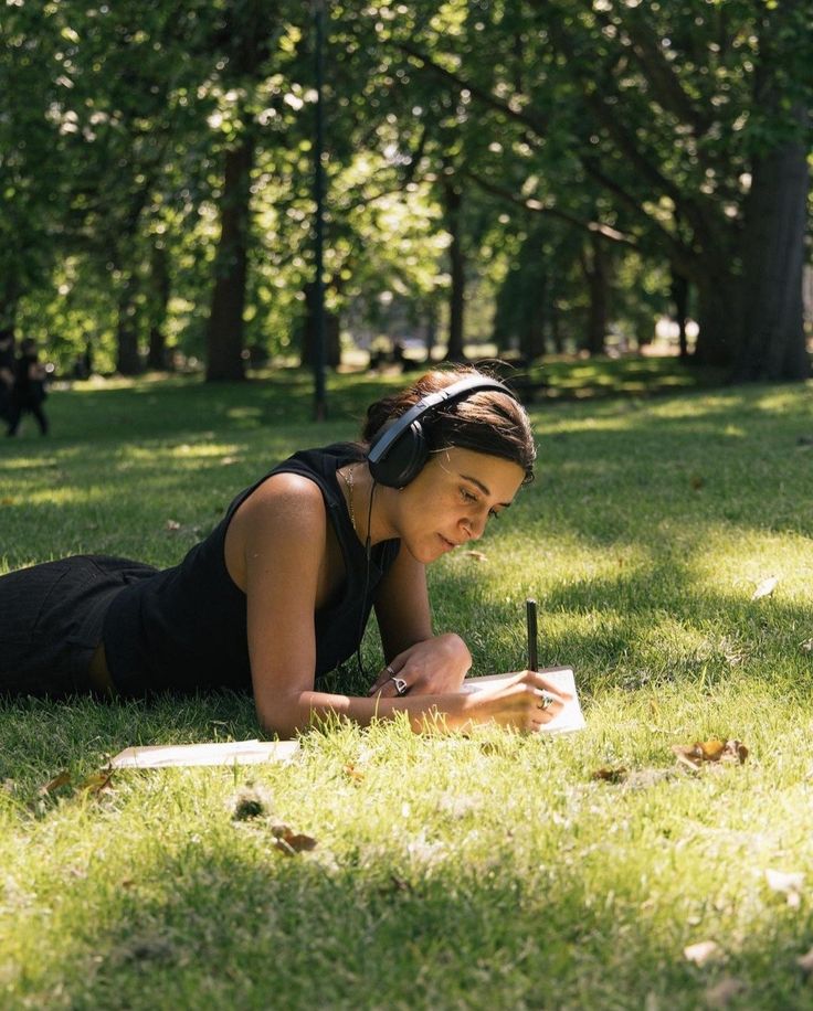 74235ac5-f86b-404e-bfb4-7288762b3dd6 Woman wearing headphones writing in a notebook while lying on grass in a sunny park. | Sky Rye Design Woman wearing headphones writing in a notebook while lying on grass in a sunny park.