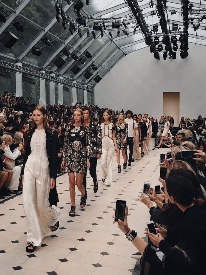 Fashion show runway with models showcasing trendy outfits, surrounded by an audience capturing photos.