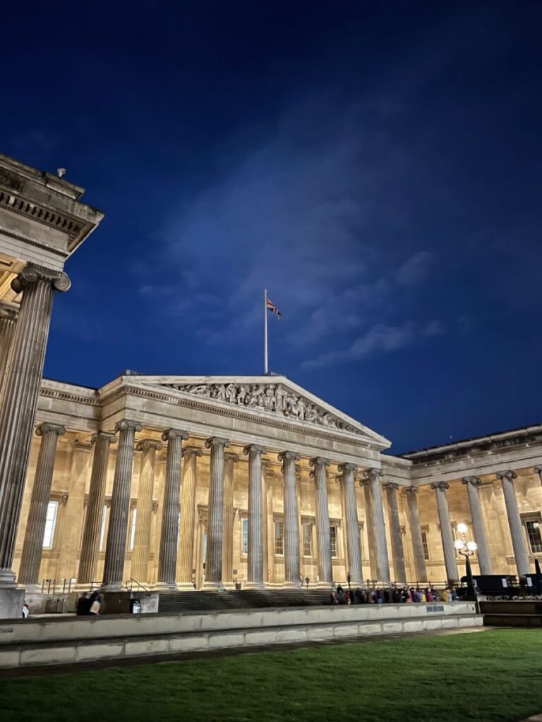 Night view of illuminated neoclassical building with columns and flag on top, against a deep blue sky.