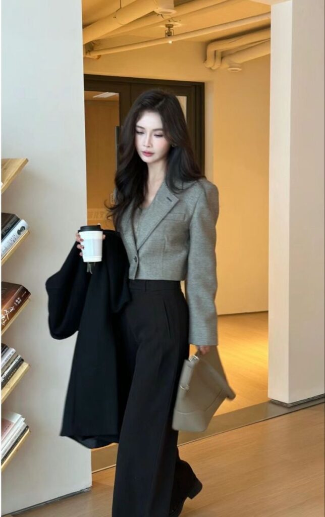 Woman in office attire holding coffee and coat, standing near a bookshelf in a modern interior.
