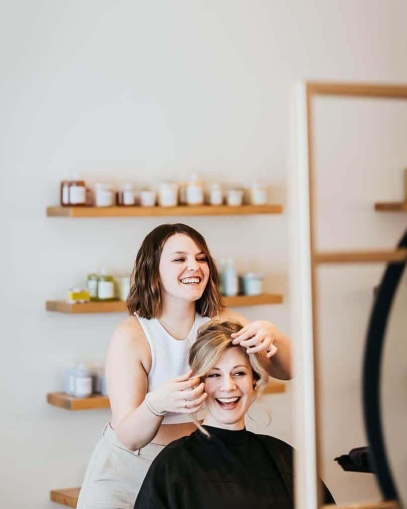 Hair stylist smiling while styling client's hair in salon. Shelves with products in background.