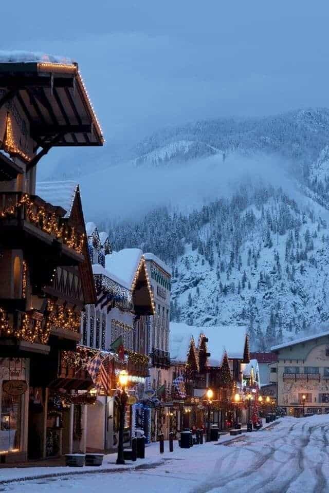 78b0f60c-fbaa-4449-947a-ee33f4bb0c08 Snow-covered village street with festive lights and alpine mountains in the background during winter twilight. | Sky Rye Design Snow-covered village street with festive lights and alpine mountains in the background during winter twilight.