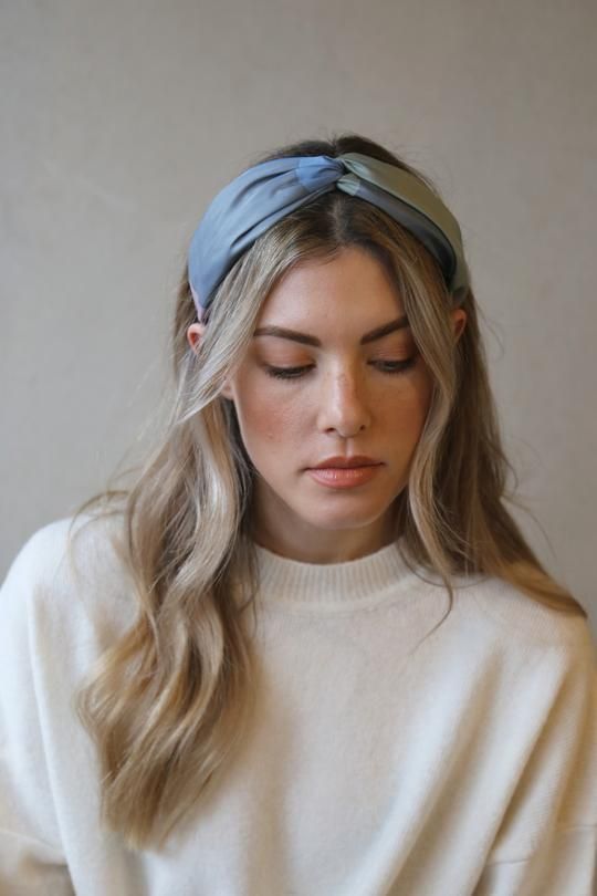 Woman with wavy blonde hair wearing a headband and white sweater, looking down thoughtfully.
