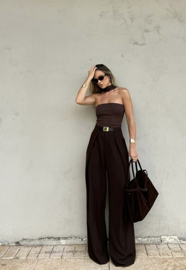 Woman in stylish brown strapless jumpsuit with sunglasses, holding a tote bag, posing against a neutral wall.
