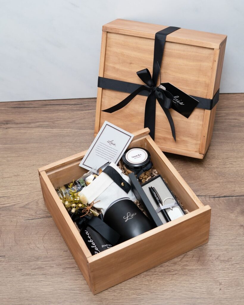 Wooden gift box with luxury items, black ribbon, and handwritten card on wooden surface, perfect for special occasions.