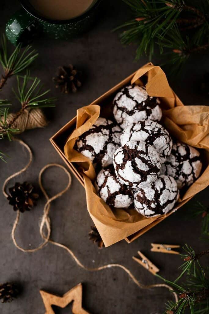 Festive chocolate crinkle cookies in a box with holiday decor, pine branches, and a cup of coffee.