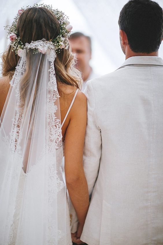Bride and groom hold hands in elegant wedding attire, featuring lace veil and floral hairpiece.