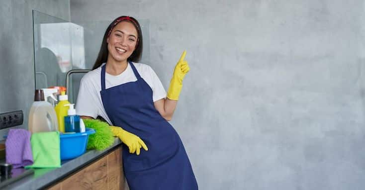 _ 8 Smiling woman in apron with cleaning supplies, pointing up in modern kitchen. Home cleaning concept. | Sky Rye Design Smiling woman in apron with cleaning supplies, pointing up in modern kitchen. Home cleaning concept.
