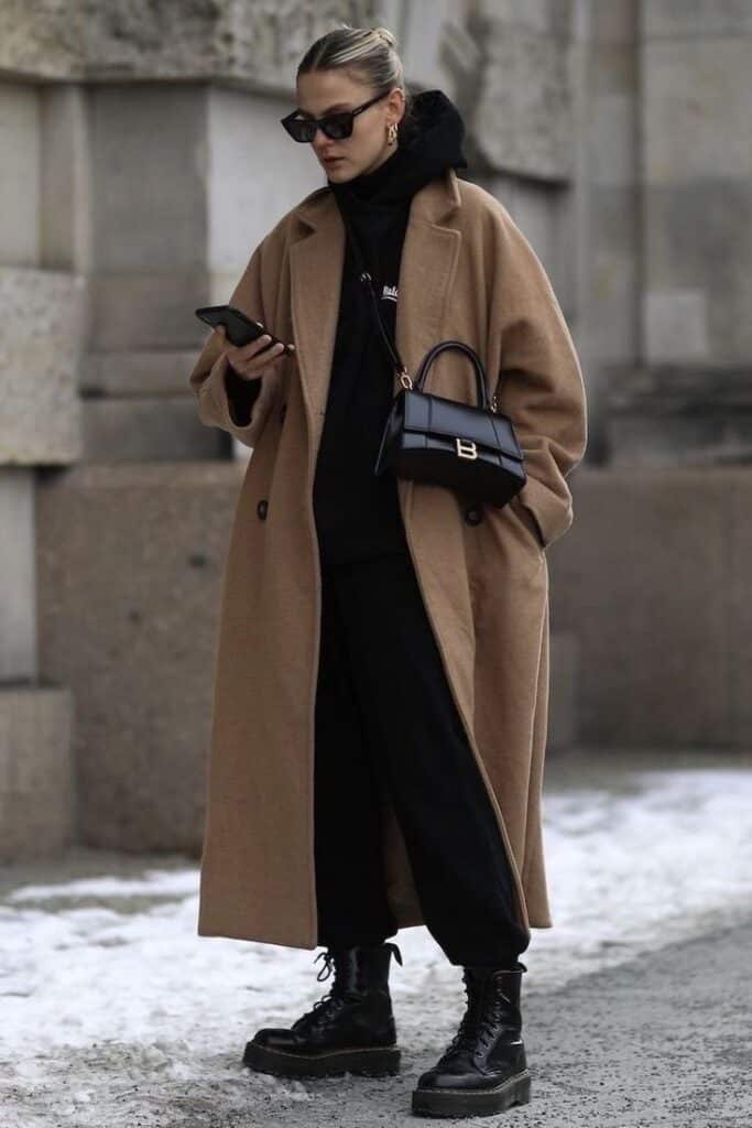 Woman in a stylish long brown coat, black outfit, holding phone and handbag, wearing sunglasses and boots.
