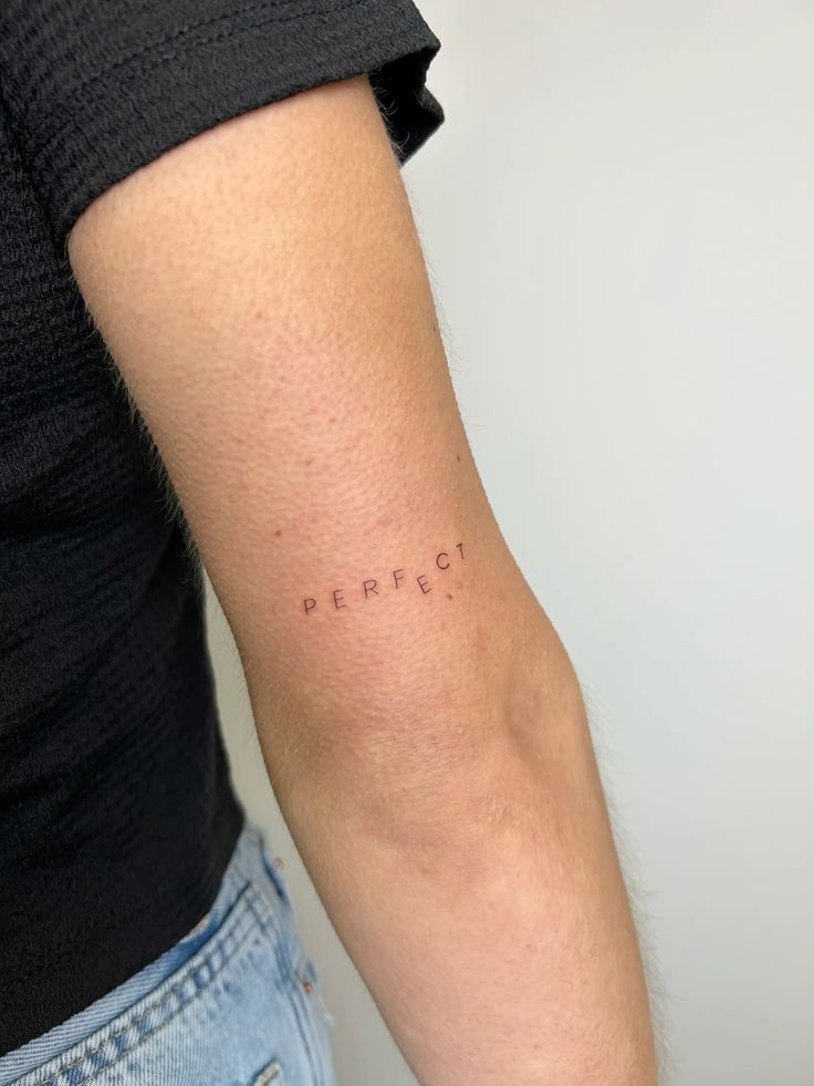 Minimalist tattoo on arm spelling PERFECT vertically aligned on skin, next to black top and jeans.