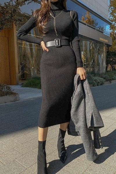 Woman in a black ribbed dress with belt, holding a gray coat outdoors. Fashionable fall street style.