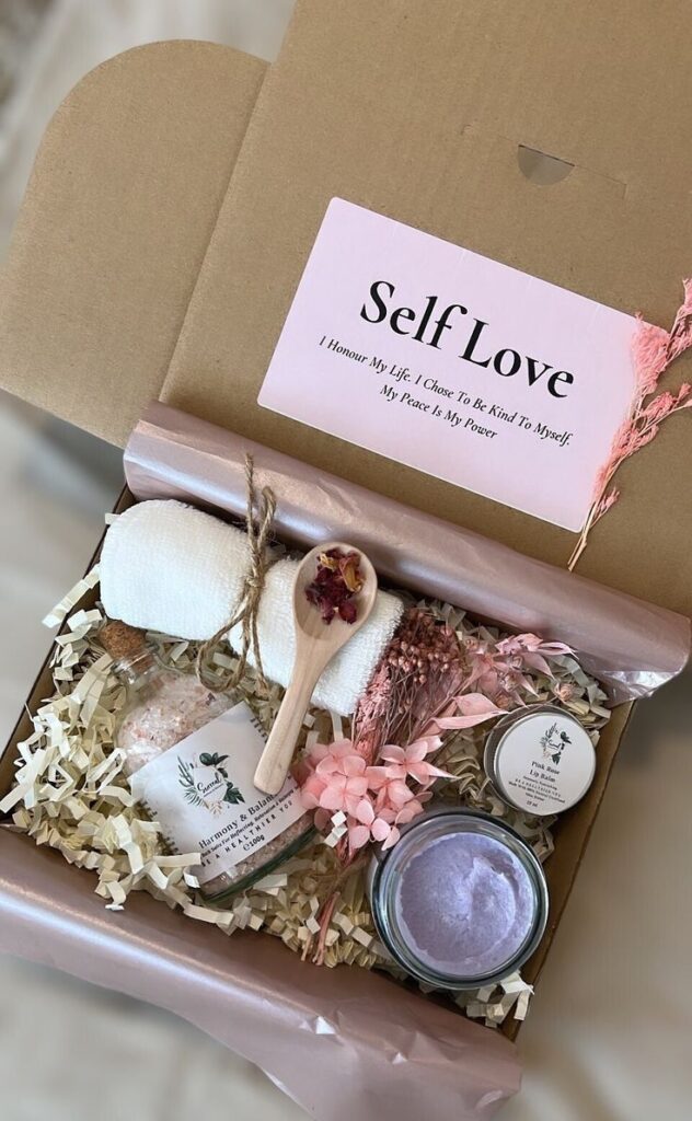 Self-care gift box with bath salts, lip balm, flowers, and towel, labeled Self Love for relaxation and pampering.