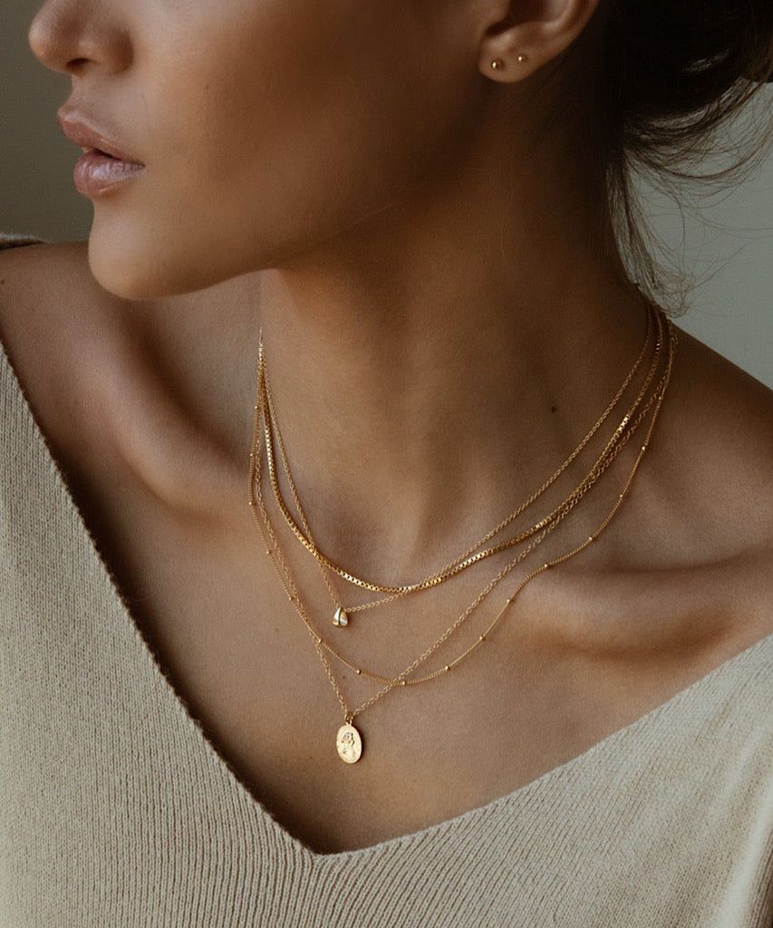 833fb1aff00e8027a08c13293ad4615c Woman wearing layered gold necklaces on a beige top, showcasing elegant jewelry style. | Sky Rye Design Woman wearing layered gold necklaces on a beige top, showcasing elegant jewelry style.