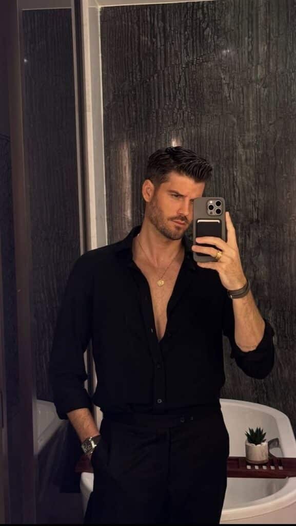 Man in a black shirt taking a mirror selfie in a sophisticated bathroom setting with a stylish pose.