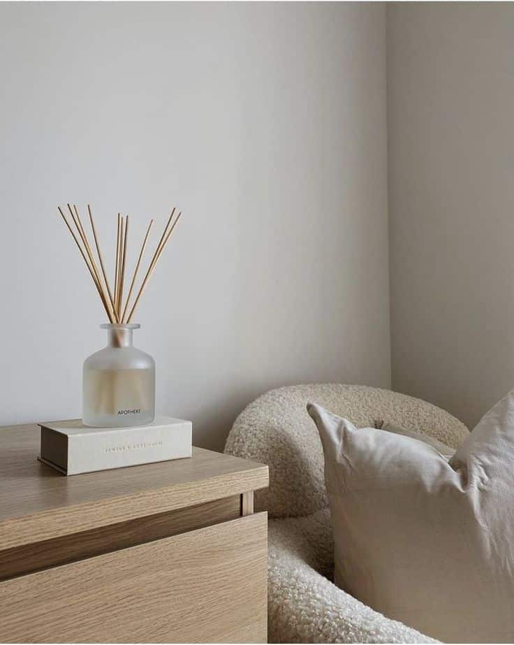 857b085a-6a49-4a79-b501-c1ca7802ea63 Minimalist cozy corner with a plush chair, pillow, wooden table, book, and reed diffuser in a bright, neutral-toned space. | Sky Rye Design Minimalist cozy corner with a plush chair, pillow, wooden table, book, and reed diffuser in a bright, neutral-toned space.