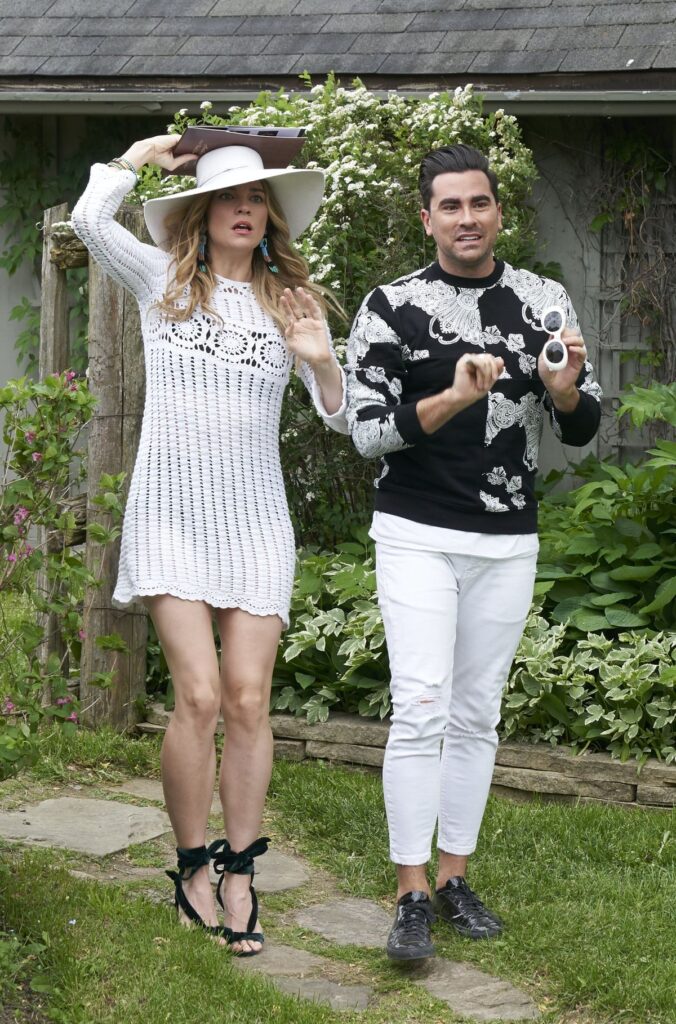 Fashionable duo outdoors; woman in white dress and hat, man in patterned sweater. Garden setting, casual style.