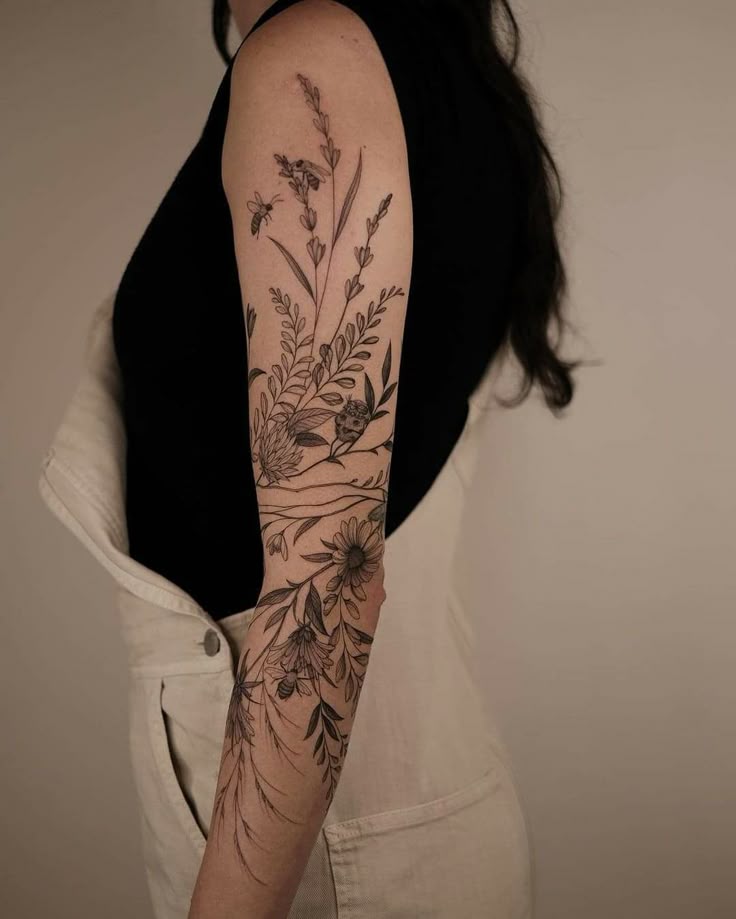 Floral arm tattoo with bees on a woman's arm wearing a black top and white overalls.