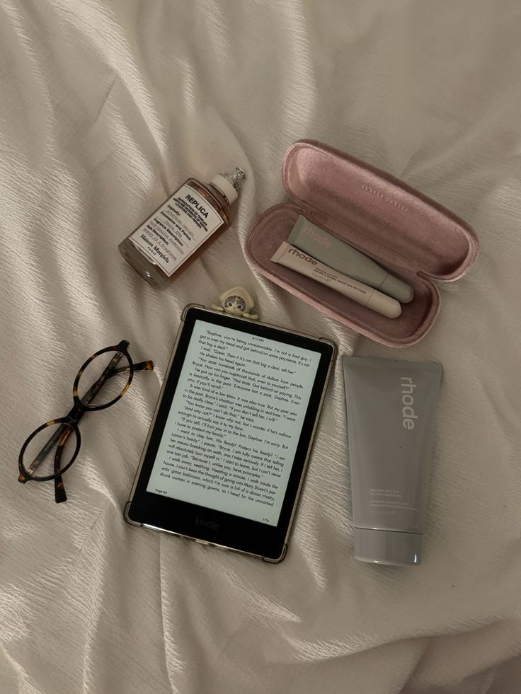 Kindle with glasses, fragrance bottle, and skincare products on a bed, creating a cozy reading and self-care vibe.