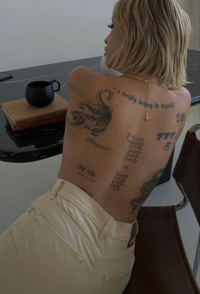 9059ac7255e78cfd2ce2f176d51dfc34 Person with back tattoos including a tiger and text, leaning on a table, next to a black mug and books. | Sky Rye Design Person with back tattoos including a tiger and text, leaning on a table, next to a black mug and books.