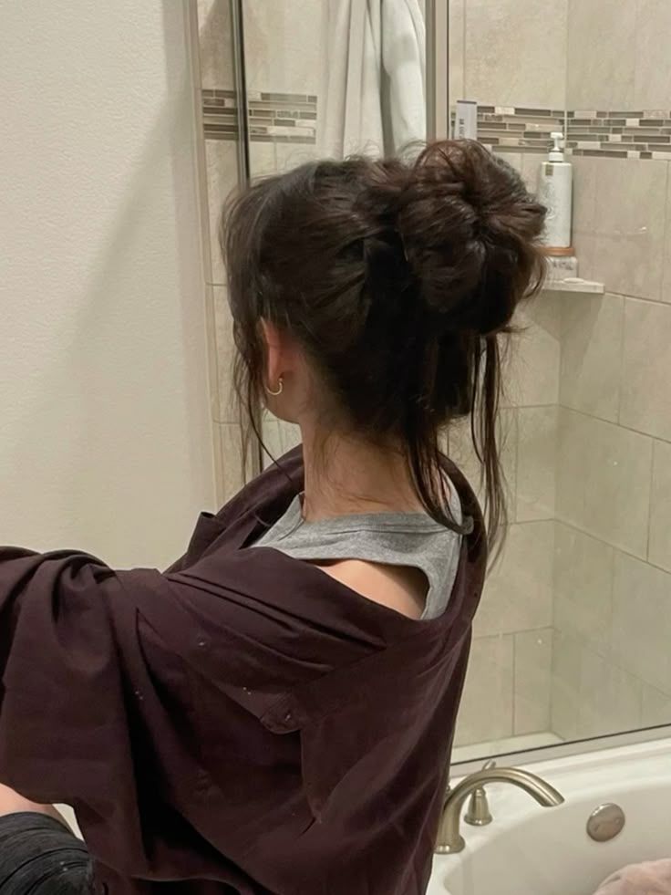 Woman in casual attire with messy bun in a bathroom setting, facing away from the camera.