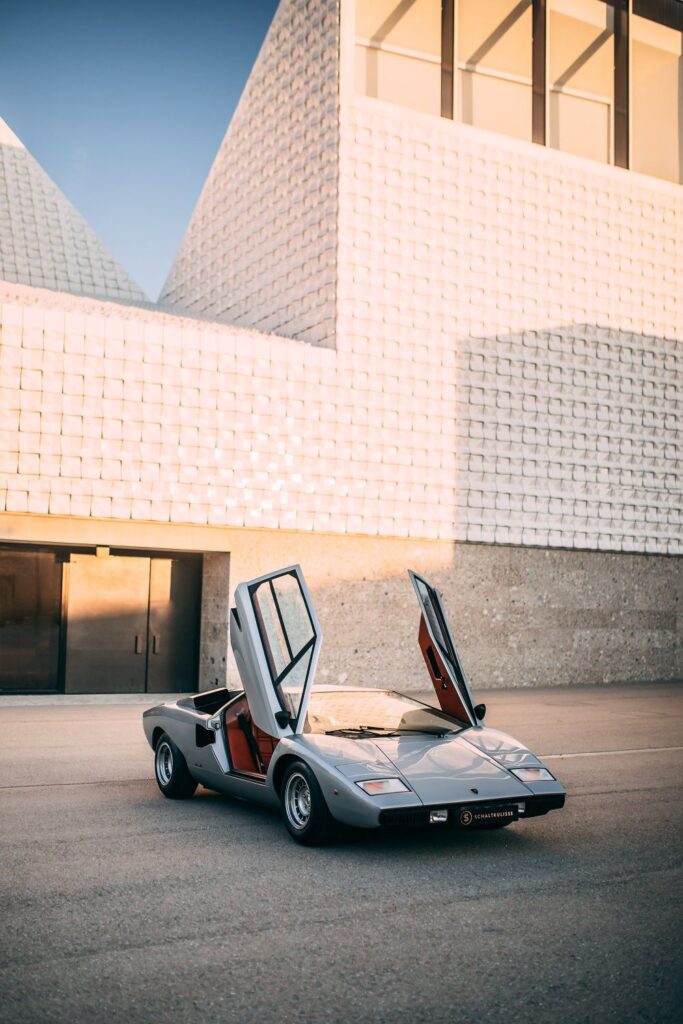 Vintage silver sports car with scissor doors open, parked in front of modern architectural building under a clear sky.