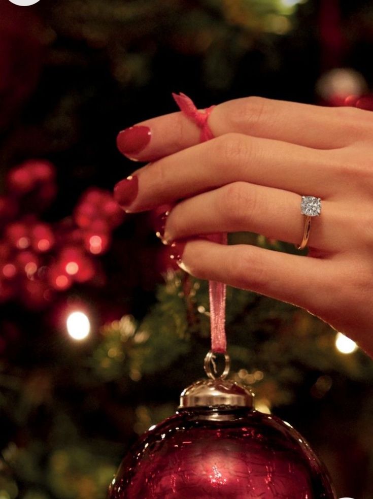 94607cdf-18ff-4ace-b06d-a848c24baa9b Hand with red nails and diamond ring holding red ornament on Christmas tree. Festive holiday decoration. | Sky Rye Design Hand with red nails and diamond ring holding red ornament on Christmas tree. Festive holiday decoration.