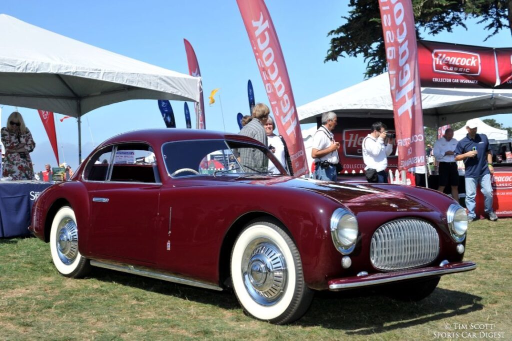 Vintage maroon Italian sports car on display at a car show with tents and banners in the background.