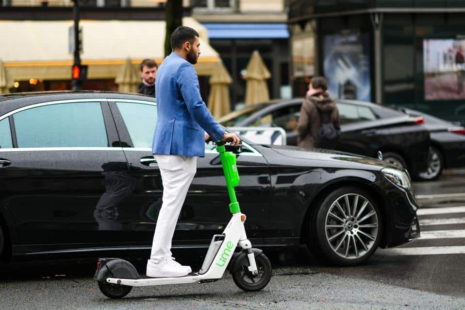 960x0 Man rides Lime e-scooter on city street in stylish blue jacket and white pants, near parked black car. | Sky Rye Design Man rides Lime e-scooter on city street in stylish blue jacket and white pants, near parked black car.