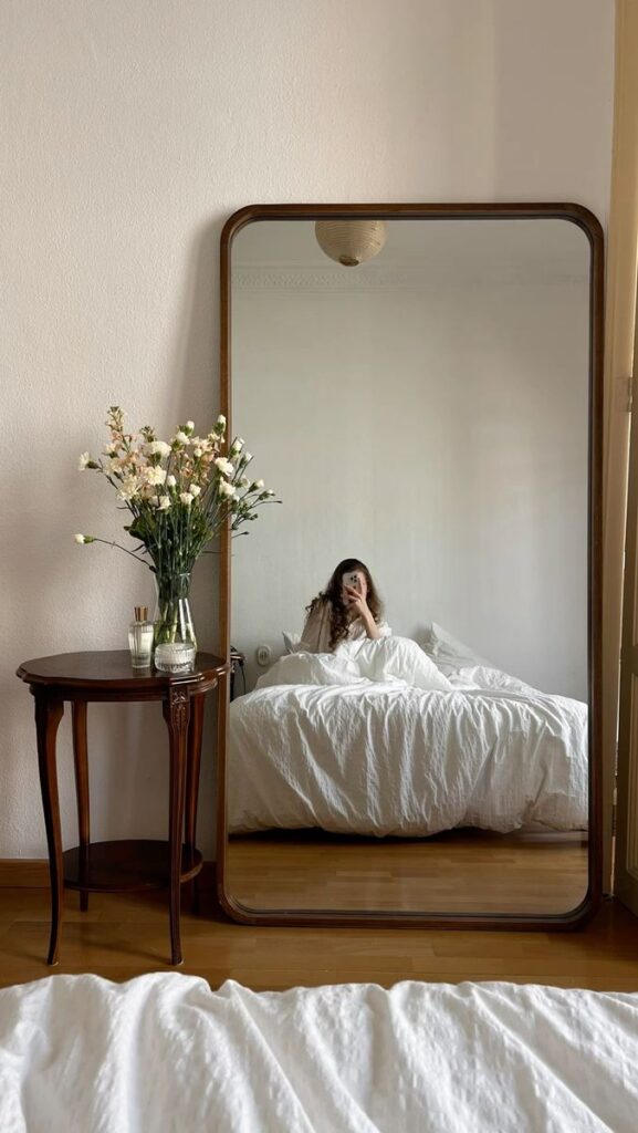 Cozy bedroom with a large mirror, white bedding, and flowers on a wooden table - minimalist decor.