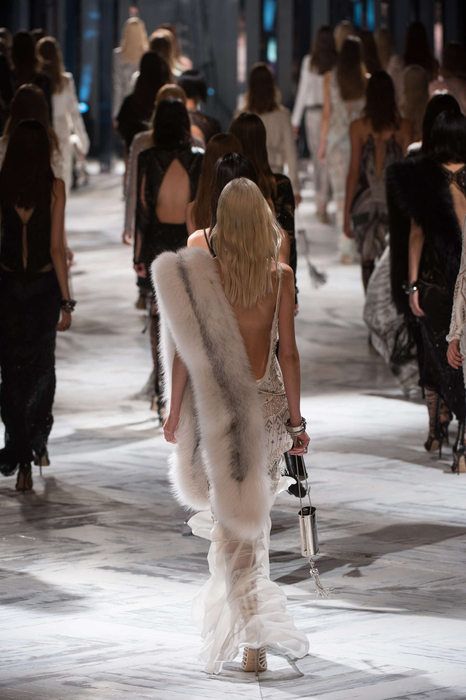Models walking down runway in elegant fashion show, wearing diverse styles including fur and intricate dresses.