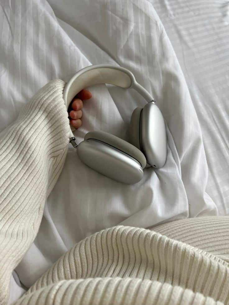 Silver headphones on a cozy, beige sweater, placed on a white bed, showcasing comfort and style.