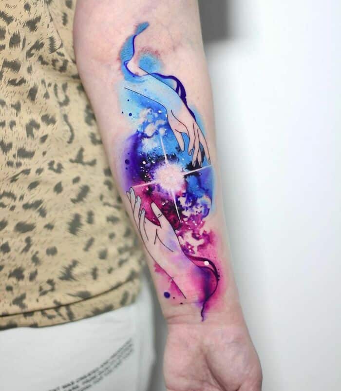 98 Watercolor Tattoos That Are Truly Ethereal Colorful watercolor tattoo of hands reaching, with a cosmic background on a person's forearm. | Sky Rye Design Colorful watercolor tattoo of hands reaching, with a cosmic background on a person's forearm.