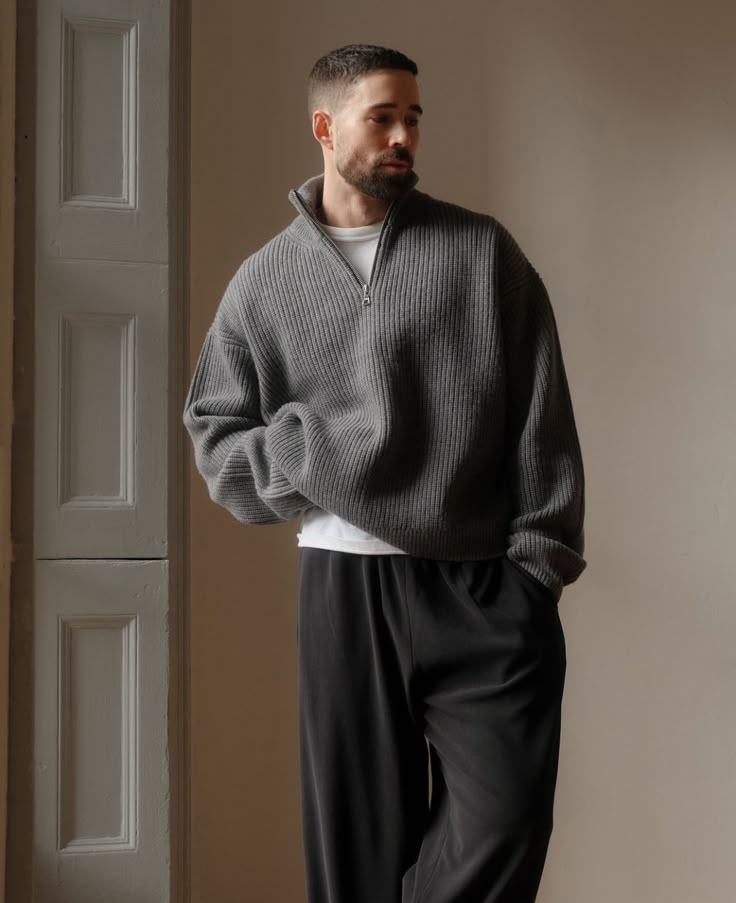 9800108f-4a44-4a03-80fa-928d9766fe9c Man wearing a gray ribbed sweater and black pants, standing in a softly lit room with a thoughtful expression. | Sky Rye Design Man wearing a gray ribbed sweater and black pants, standing in a softly lit room with a thoughtful expression.