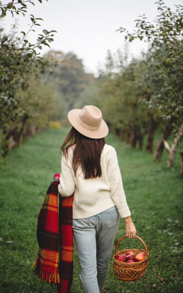 Woman in sweater and hat walks through orchard carrying apples and a plaid blanket, enjoying autumn outdoors.