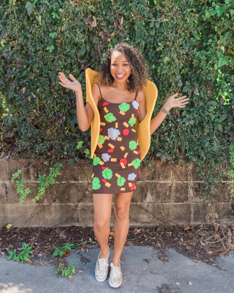 Woman in a vibrant taco costume smiling, with a lush green leafy wall background.