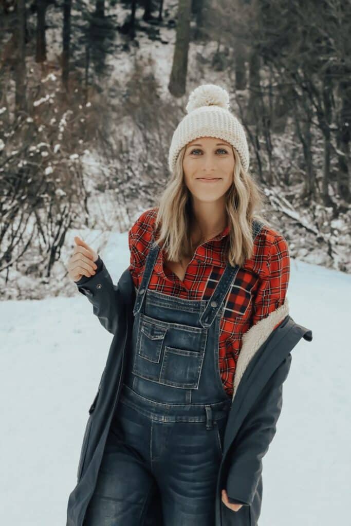 Woman in a red plaid shirt and overalls, wearing a winter hat, stands smiling in a snowy forest path.