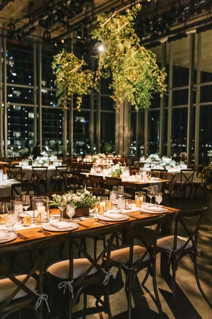 Elegant indoor event setup with candlelit tables, floral centerpieces, and hanging greenery, against a city backdrop.