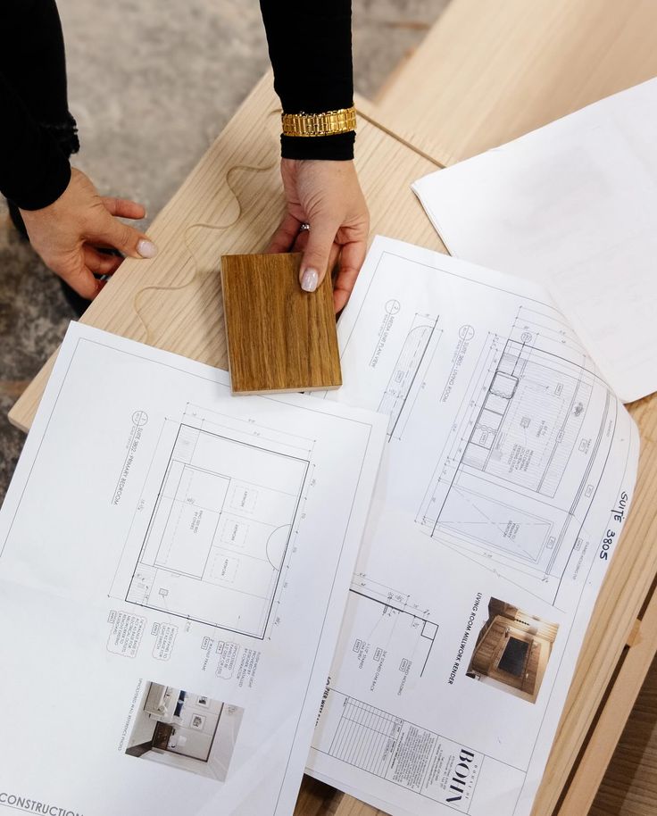 A stop in at one of our custom furniture and millwork collaborators to check up on progress for an upcoming project_ Person reviewing architectural blueprints with a wooden sample for interior design decisions. | Sky Rye Design Person reviewing architectural blueprints with a wooden sample for interior design decisions.