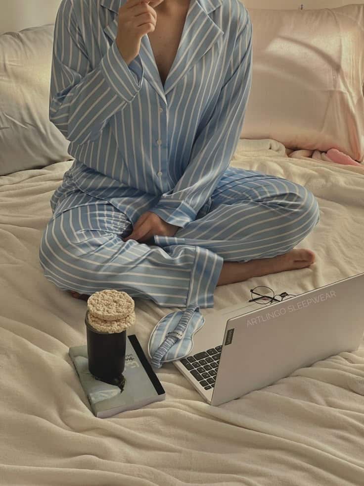 Person in striped pajamas working on a laptop with coffee and rice cakes on a bed. Cozy and relaxed work from home.