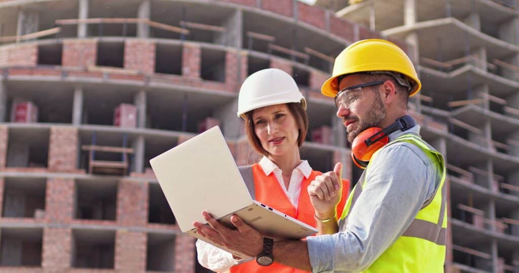 Builders foreman and female architect standing in front of construction site and look at the lap top discussing their job The concept of new technologies in construction Front view Close up Construction workers discussing plans on a laptop at a building site, wearing hard hats and safety gear. | Sky Rye Design Construction workers discussing plans on a laptop at a building site, wearing hard hats and safety gear.