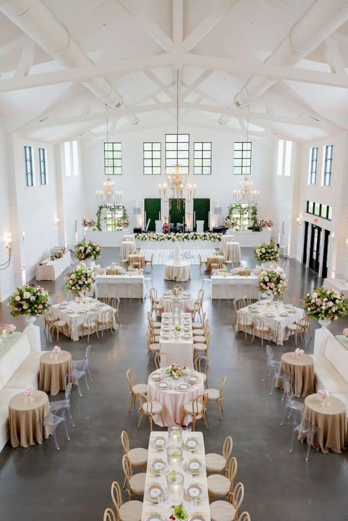 Elegant wedding reception in a bright hall with chandeliers, floral centerpieces, and neatly arranged tables.