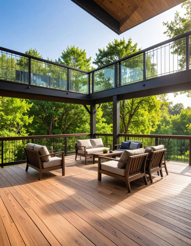 Modern wooden deck with outdoor furniture and glass railings, surrounded by lush green trees under a clear blue sky.
