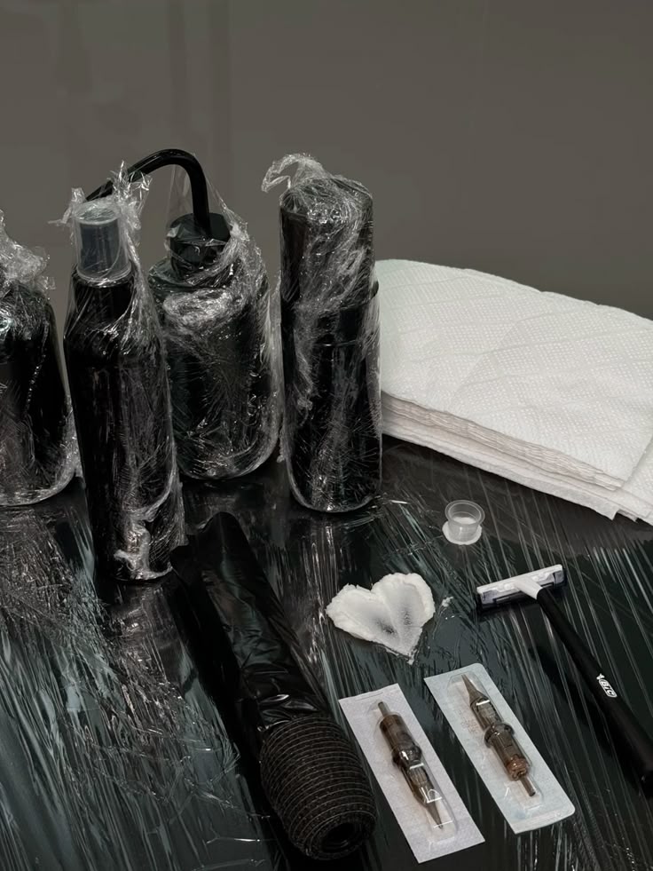 Aesthetic Tattoo setup: wrapped bottles, paper towels, ink caps, razor, and needles on a sterile black surface. | Sky Rye Design Tattoo setup: wrapped bottles, paper towels, ink caps, razor, and needles on a sterile black surface.