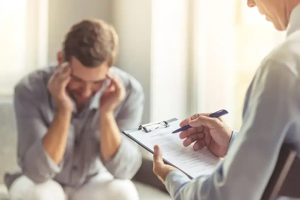 Therapist counseling distressed man, holding clipboard in bright office setting.