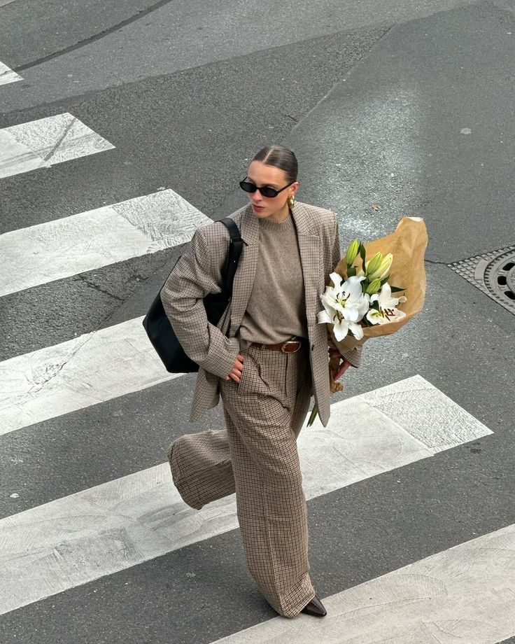 Stylish woman in plaid suit holding white lilies, crosses street with confidence. Fashionable urban scene.